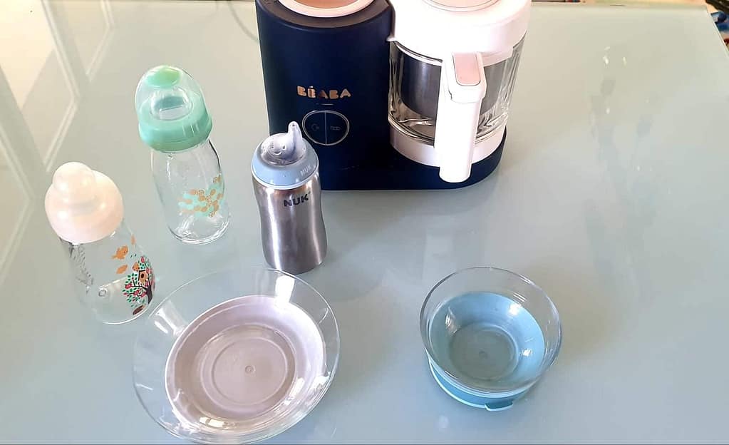 Une photo montrant des biberons en verre, un biberon en inox, des bols en verre et un robot de cuisine pour bébé de la marque Béaba sur une table en verre.