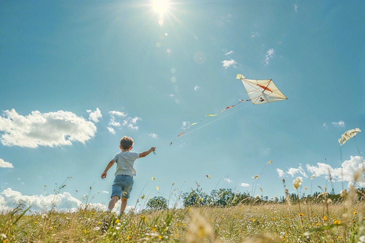 Regarder le monde à travers les yeux d'un enfant. Un jeune garçon en t-shirt blanc et short bleu fait voler un cerf-volant dans un champ ensoleillé, reflétant la simplicité et la joie de l’enfance.