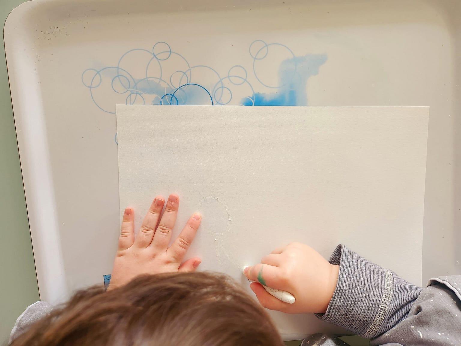 Enfant dessinant à la craie grasse blanche sur une feuille blanche, première étape de l’activité de peinture magique
