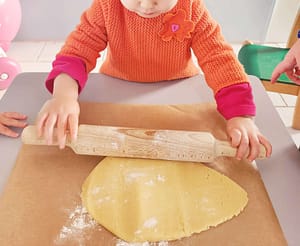 Un enfant roule la pâte à sablés de Noël avec un rouleau à pâtisserie en bois, sous la supervision d’un adulte.