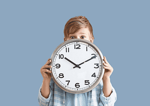 Enfant tenant une horloge devant son visage sur fond pastel, illustrant le respect du rythme de l’enfant