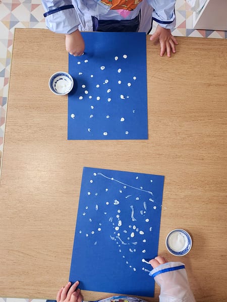 Enfants utilisant des cotons-tiges trempés dans de la gouache blanche pour dessiner des flocons de neige sur une feuille bleue