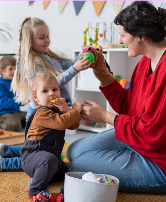 Assistante maternelle assise au sol avec deux enfants, jouant avec des jouets et une marionnette.